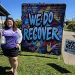 Jennifer holding candle 🕯 from the 2nd annual event in front of the "We do Recover" sign.