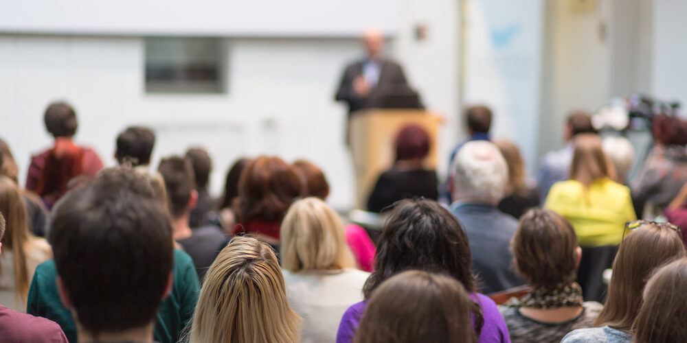 Male speaker giving presentation in lecture hall at university workshop. Audience in conference hall. Rear view of unrecognized participant in audience. Scientific conference event.